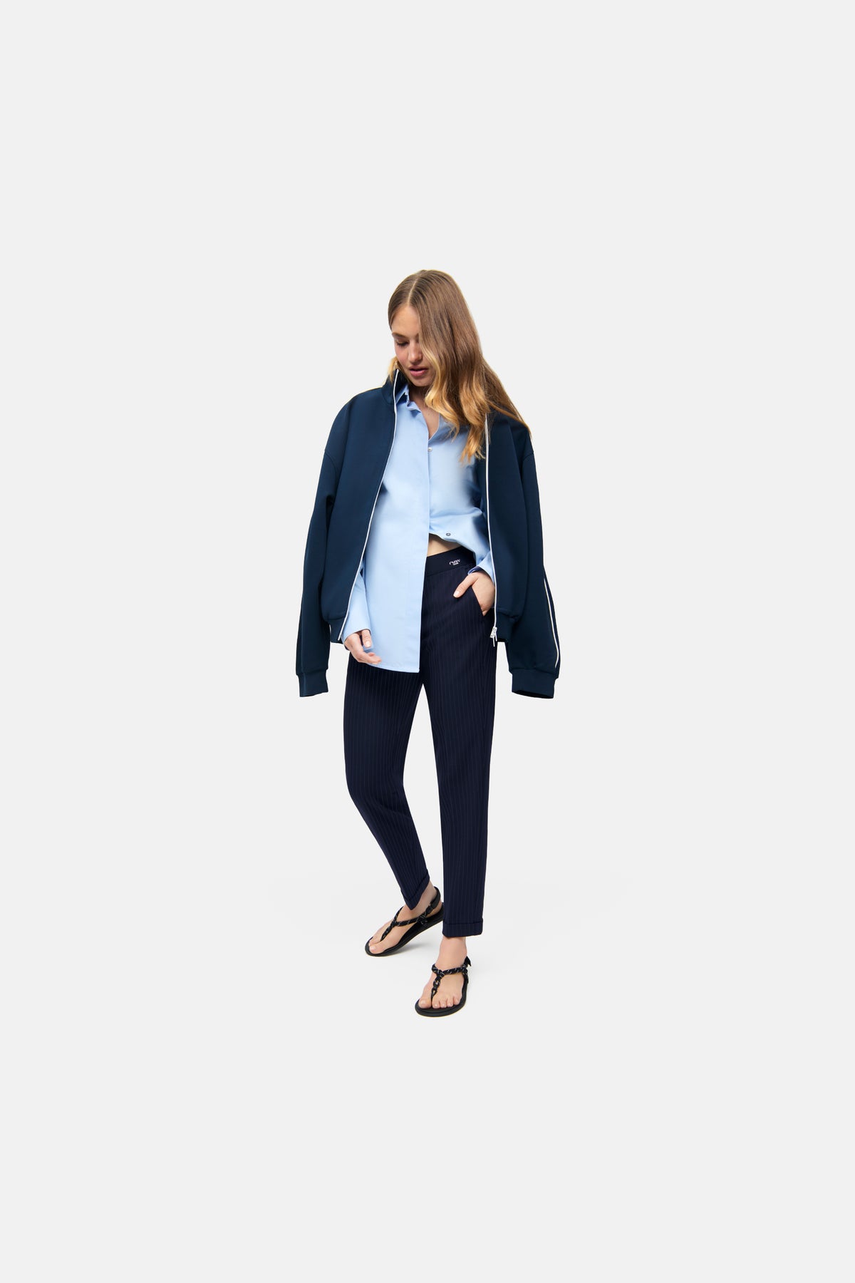 Dunkelblau gestreifte Karottenhose aus 62% Polyester, 26% Viskose, 9% Wolle und 3% Elasthan. Frau trägt die Hose mit Hemd und Jacke.