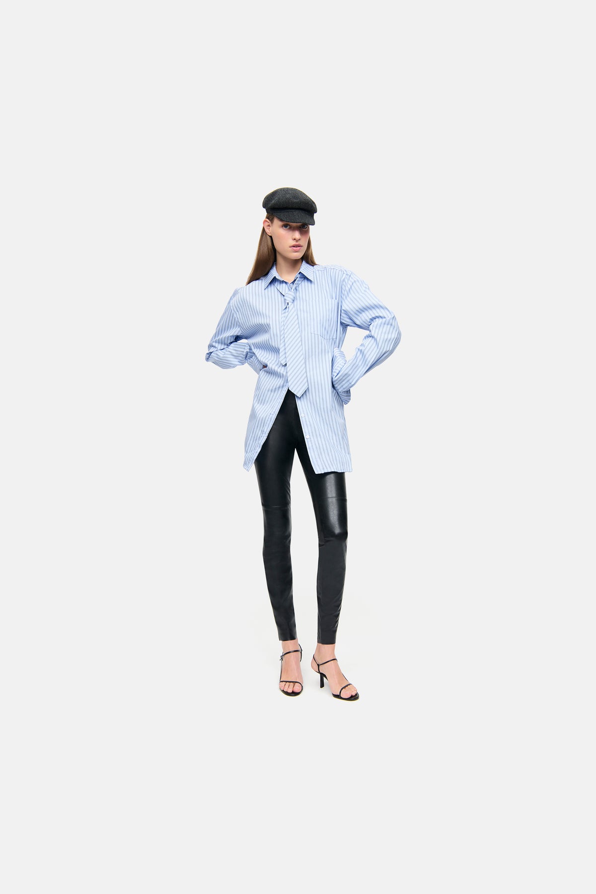 Die abgebildete Skinny-Fit Hose ist in Schwarz gehalten und besteht aus veganem Nappa mit einem elastischen Bund. Sie wird von einer Person getragen, die ein gestreiftes Hemd und Sandalen mit Absatz kombiniert hat.