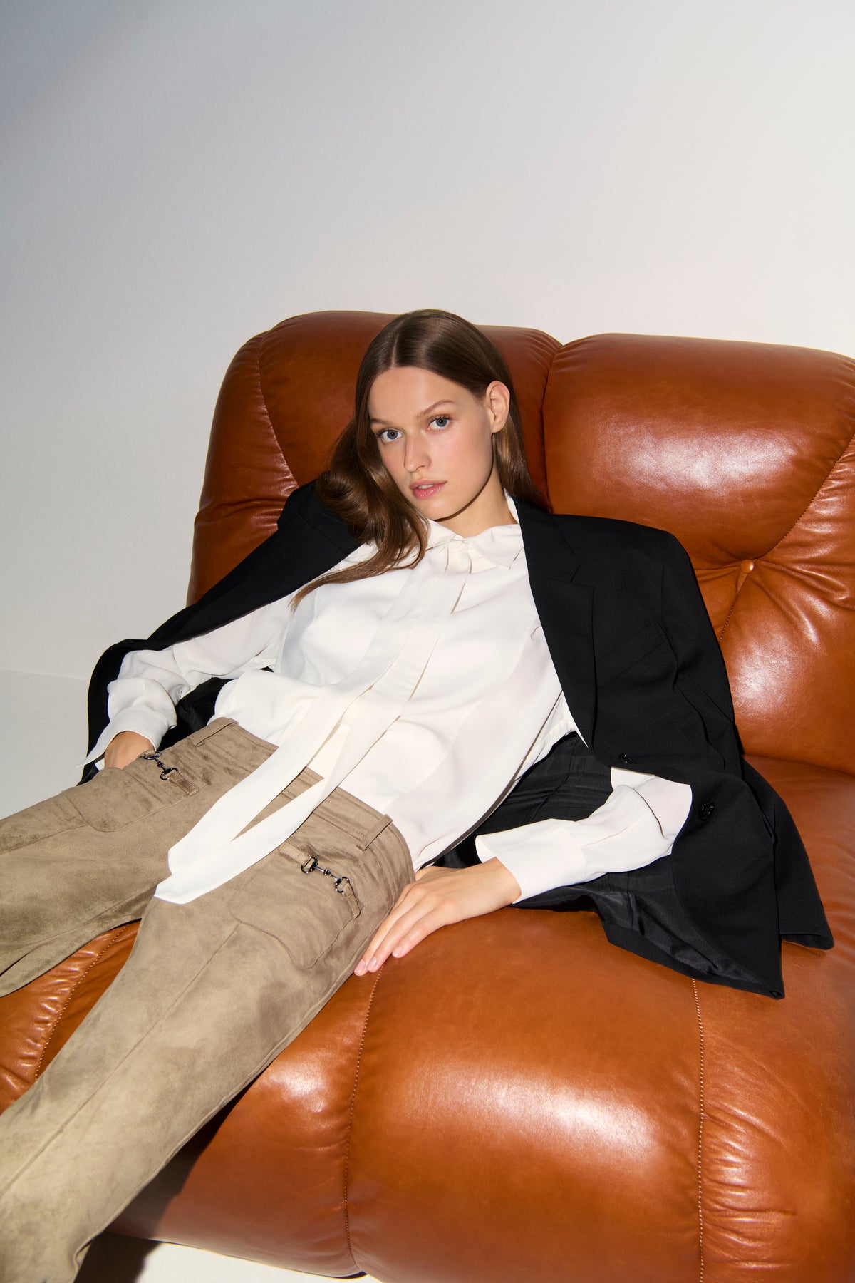 Frau in modernem Setting sitzt auf Ledersessel, trägt braune Flared-Hose aus veganem Veloursleder, 92% Polyester, 8% Elasthan.