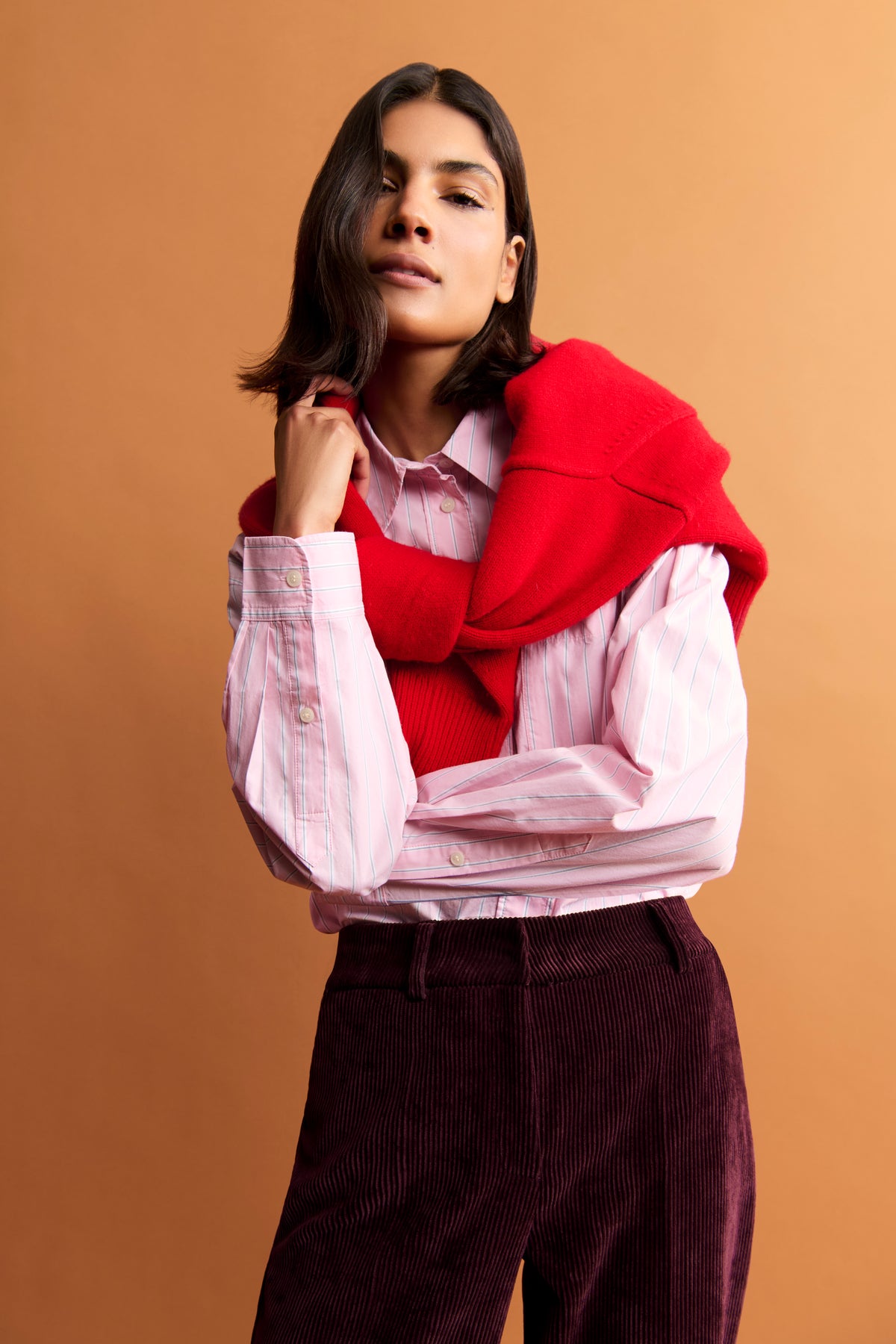 Bordeauxfarbene Cordhose im Wide-Leg-Stil, getragen mit rotem Pullover um die Schultern und rosa gestreiftem Hemd.