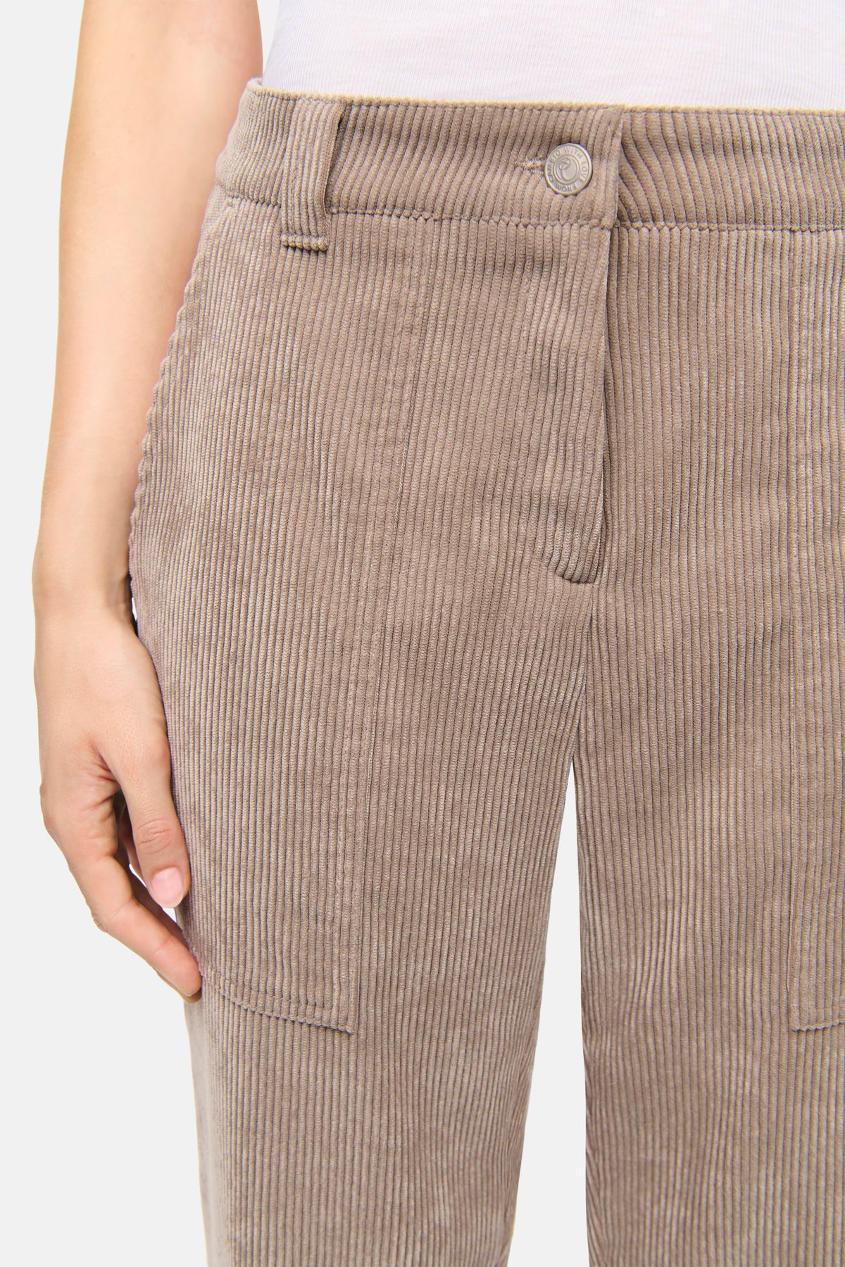 Braune Cordhose in verkürzter Karotten-Silhouette aus 90% Polyester, 8% Polyamid, 2% Elasthan. Szene zeigt Hand in Hosentasche.