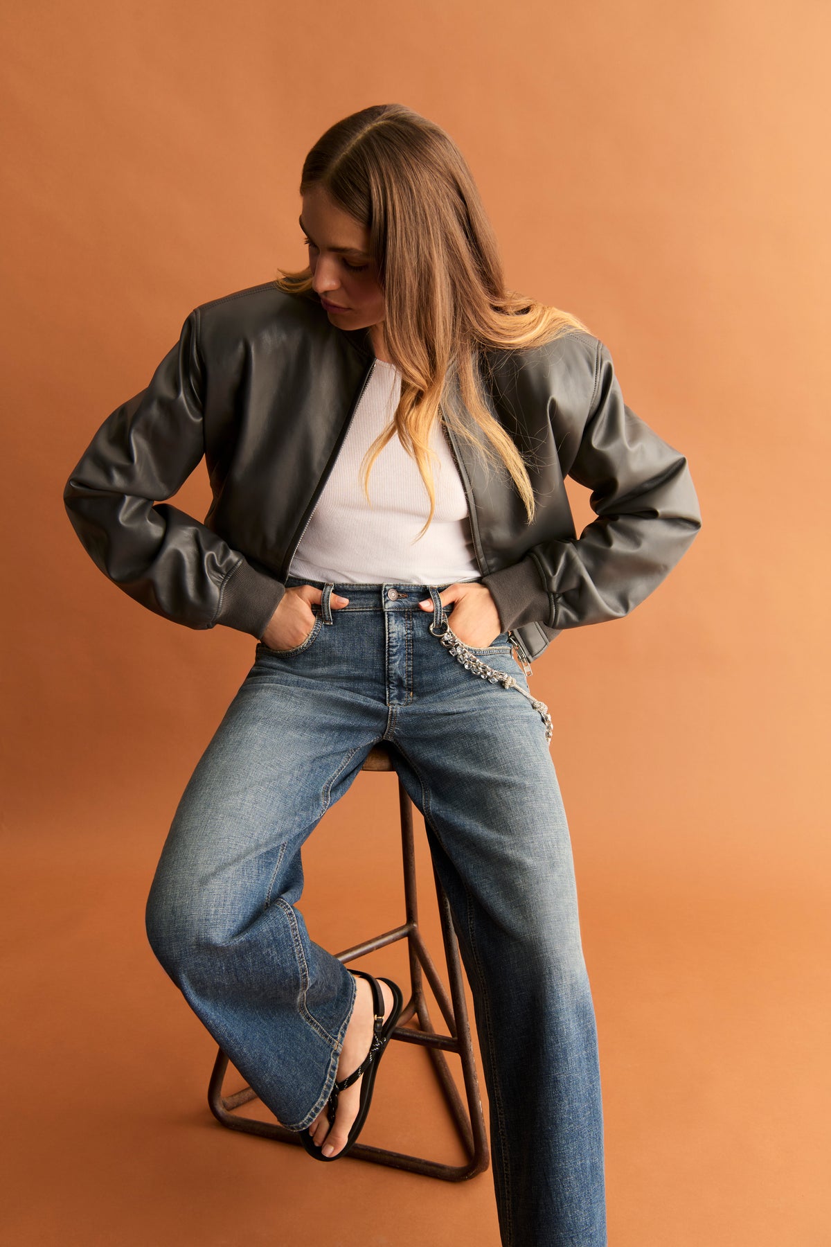 Damen trägt eine weite blaue Jeans aus 98% Baumwolle und 2% Elasthan, kombiniert mit einer Lederjacke. Sitzend auf einem Hocker.